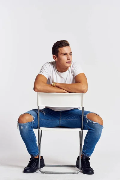 A man in jeans sits on a chair backwards and and ripped jeans T-shirt front view — Stock Photo, Image