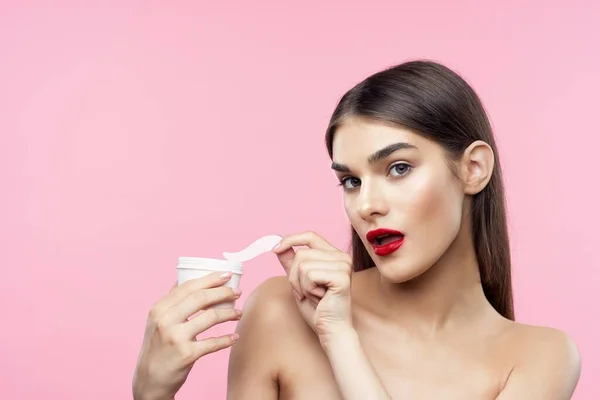 Pretty woman with bare shoulders cream in hands clear skin pink background — Stockfoto