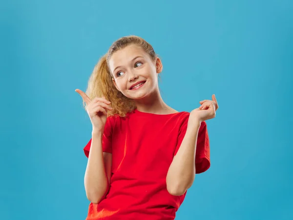 Χαρούμενο κορίτσι σε ένα κόκκινο t-shirt με σγουρά μαλλιά χειρονομίες με τα χέρια της μπλε φόντο — Φωτογραφία Αρχείου