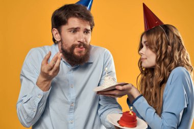 Sarı arka planda ellerinde pastayla doğum günü partisi veren erkek ve kadın.