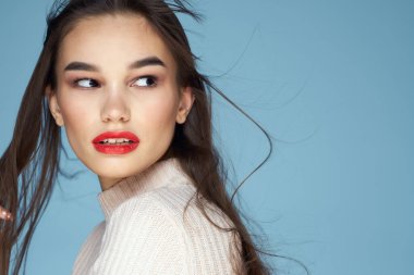 Beyaz kazaklı esmer, kırmızı dudaklı, uzun saçlı, mavi arkaplan.