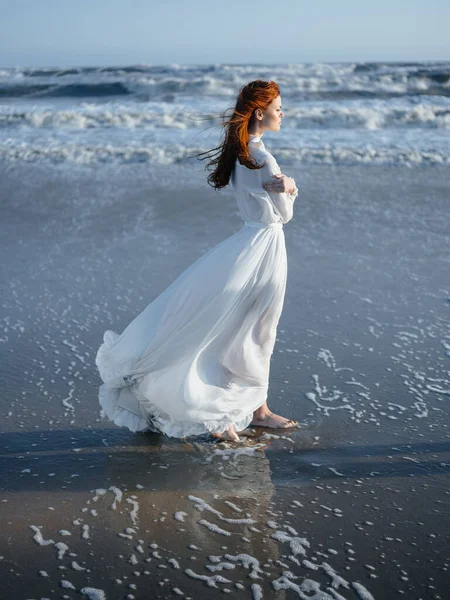 Μια γυναίκα με λευκό φόρεμα περπατάει στην υγρή άμμο στην ακτή του ωκεανού σε πλήρη ανάπτυξη. — Φωτογραφία Αρχείου