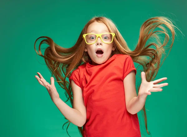 Menina em uma camiseta vermelha com emoções de cabelo solto óculos amarelos Fundo verde — Fotografia de Stock