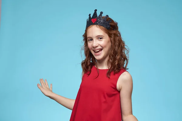 Dívka s kudrnaté vlasy s korunou na hlavě červené šaty životní styl modré pozadí — Stock fotografie