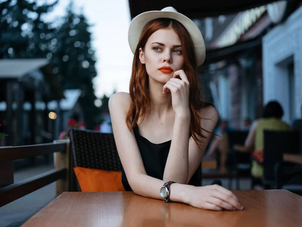 Une femme avec un chapeau blanc et une robe noire à une table dans un café dans la rue — Photo