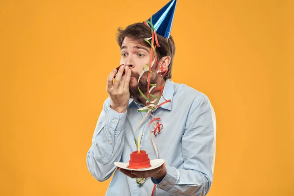 Hombre alegre con un pastel en un fondo amarillo cumpleaños días festivos gorra en su cabeza —  Fotos de Stock