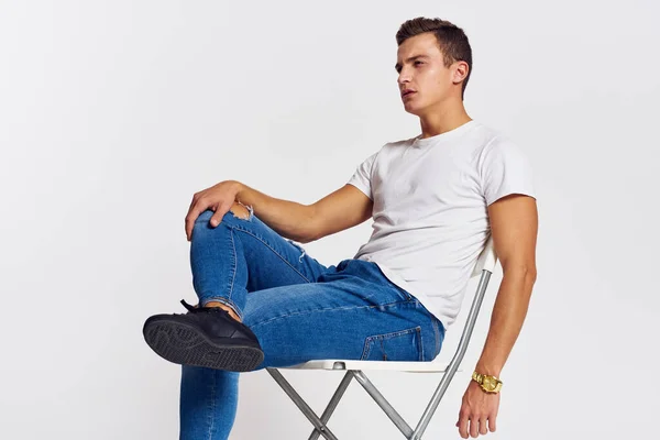 Man on a chair indoors torn jeans white t-shirt handsome face model light background — Stock Photo, Image