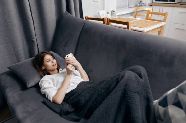 Kadın kanepeye uzanıyor ve telefon ekranının üst görüntüsüne bakıyor.