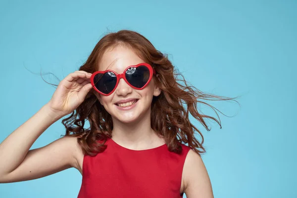 Dziewczynka w okularach przeciwsłonecznych w postaci serc czerwona sukienka kręcone włosy niebieskie tło — Zdjęcie stockowe