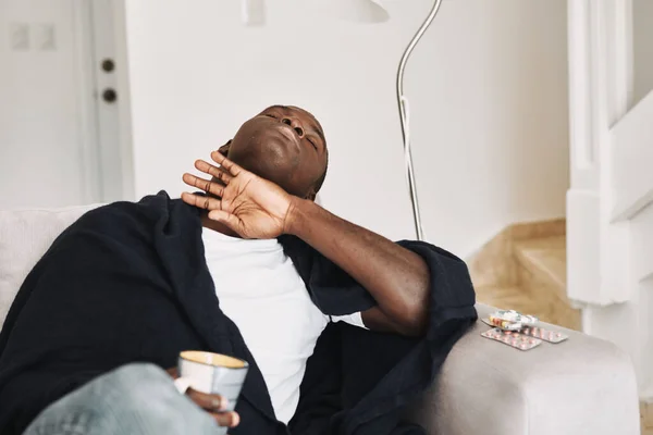Człowiek o afrykańskim wyglądzie zasnął na kanapie z kubkiem w ręku i tabletem na boku. — Zdjęcie stockowe