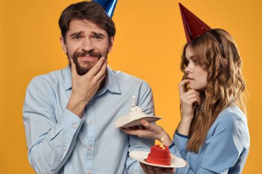 Sarı arka planda pastası olan şapkalı kadın ve erkek doğum günü partisi.