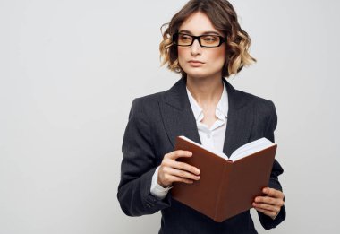 Klasik takım elbiseli bir kadın elinde bir kitapla arka planda ve gözlüklü bir kadın.