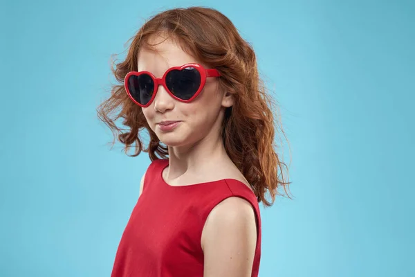 Маленькая девочка в солнечных очках в виде сердец красное платье вьющиеся волосы синий фон — стоковое фото