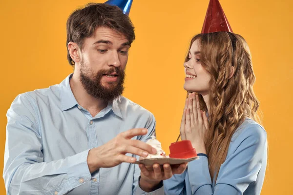 Sarı arka planda pastası olan şapkalı kadın ve erkek doğum günü partisi. — Stok fotoğraf