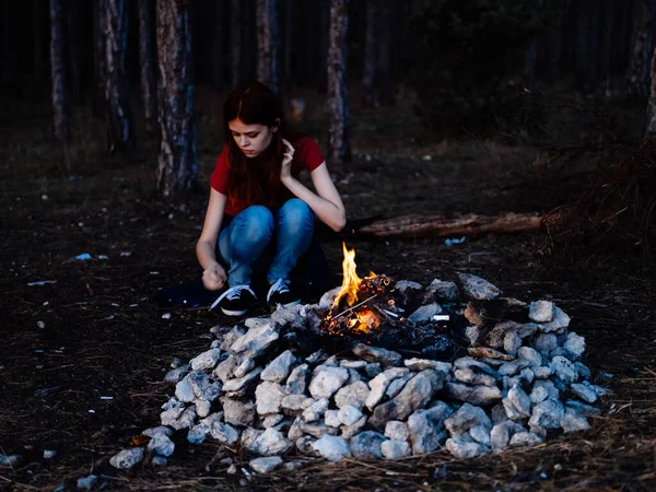 Een romantische vrouw in jeans en een T-shirt zit bij een kampvuur in de natuur — Stockfoto