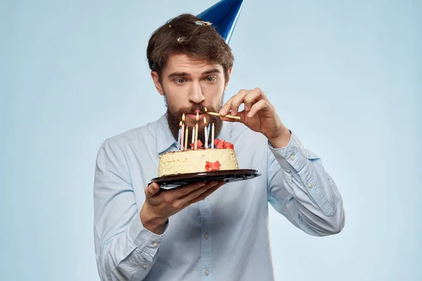 青い背景にケーキのプレートを持つ髭の男と彼の頭の上に誕生日パーティーの帽子 — ストック写真