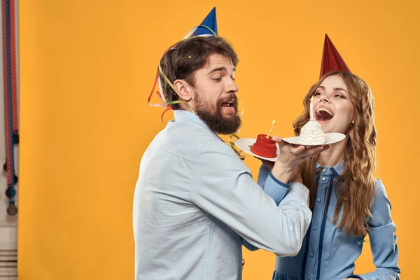 Man and woman at a party in caps and with tinsel cake fun yellow background — Stock Photo, Image