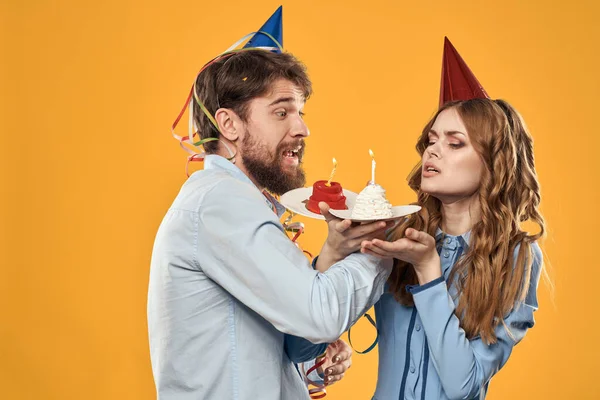 Homem e mulher em uma festa em bonés e com bolo de ouropel divertido fundo amarelo — Fotografia de Stock