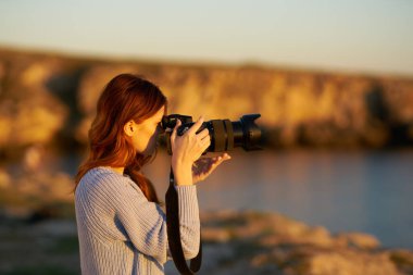 Gün batımında elinde kamera olan kadın deniz kenarındaki dağlarda.