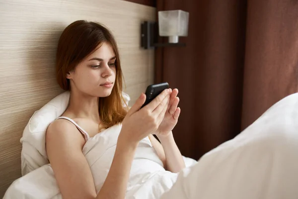 Mulher com telefone móvel encontra-se na cama perto da janela e cortinas design — Fotografia de Stock