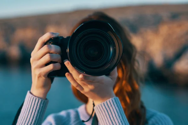 Γυναίκα κρατώντας μια φωτογραφική μηχανή με ένα φακό στη φύση στα βουνά και τη θάλασσα στο παρασκήνιο — Φωτογραφία Αρχείου