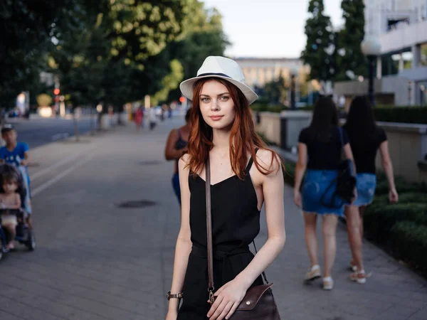 Mulher na moda em um vestido preto e em um chapéu branco caminha na rua no parque — Fotografia de Stock