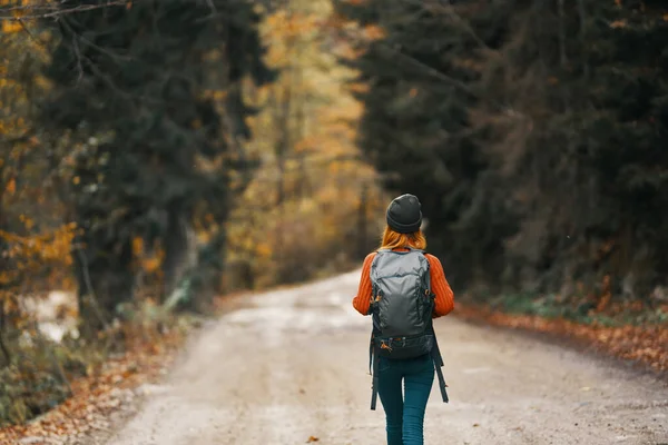 Mujer con una mochila en un sombrero y un suéter naranja en la carretera en el bosque de otoño — Foto de Stock