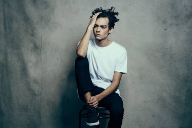 Beyaz tişörtlü adam sandalyeye oturur. Kıvırcık saç stili.