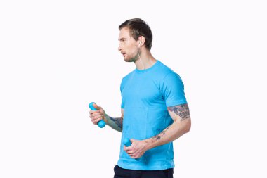 Hafif arka planda dambılları olan mavi tişörtlü genç adam ve siyah pantolon fitness modeli.