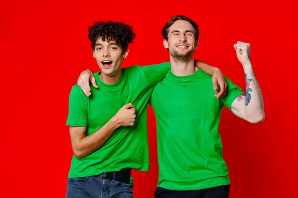 Dos amigos alegres abrazan verde camisetas emociones comunicación fondo rojo — Foto de Stock