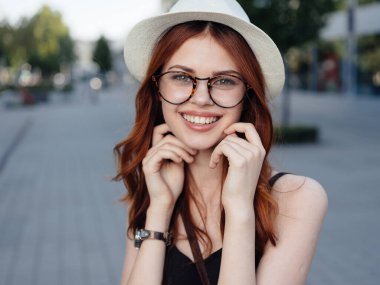 Beyaz şapkalı, gözlüklü, kızıl saçlı bir kadın yazın sokakta yürüyor.