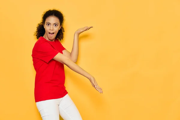 Mooie vrouw met krullend haar in een rood t-shirt op een gele achtergrond gebaar met haar handen — Stockfoto