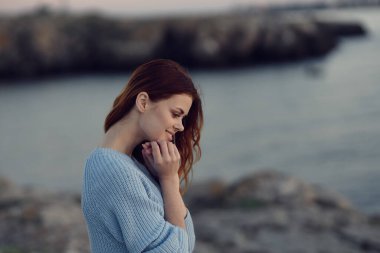 Doğada nehrin kenarında mavi kazaklı kızıl saçlı bir kadın.