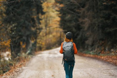 Sırt çantalı, şapkalı ve turuncu kazaklı bir kadın sonbahar ormanında yolda.