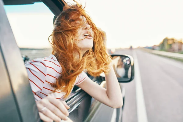Mujer pelirroja feliz mira por la ventana del coche y conduce en la carretera, pista vacaciones de verano — Foto de Stock