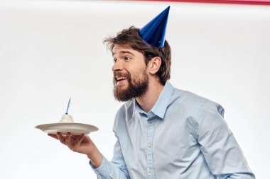  Elinde pasta tabağı ve açık arkaplanda mavi gömlekli bir adam doğum günü partisi şapkası takıyor.