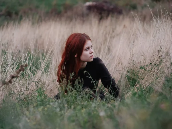 Vörös hajú nő fekete ruhában fekszik az erdőben száraz füvön a természetben — Stock Fotó