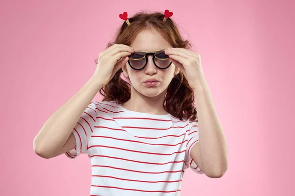 Petite fille dans des lunettes et des pinces à linge à la mode sur ses cheveux émotions fond rose coeurs — Photo