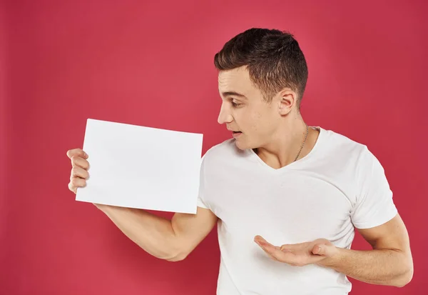 Een man met een flyer in zijn handen op een rode achtergrond in een wit t-shirt bijgesneden uitzicht — Stockfoto