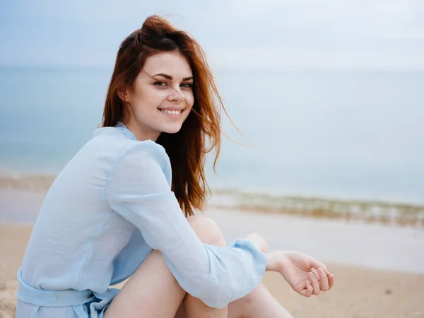 Una donna romantica in un abito blu trasparente siede sulla sabbia vicino al mare e sorride — Foto Stock