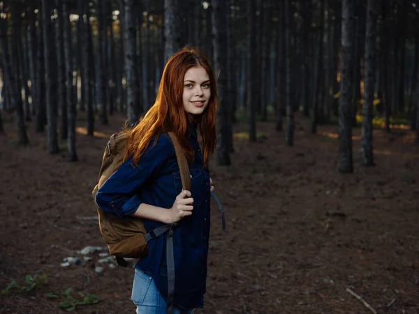 Mujer pelirroja con una camisa azul en un bosque de pinos con una mochila en la espalda —  Fotos de Stock