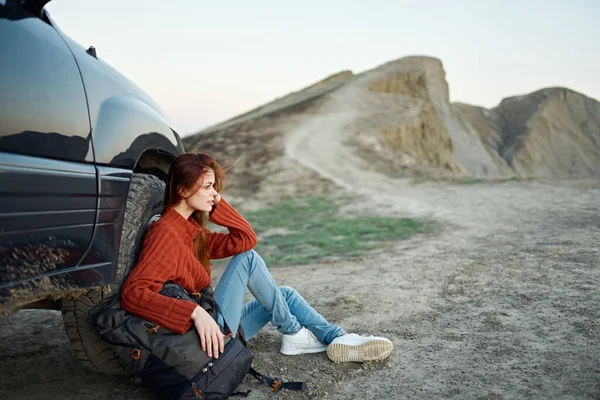 Um viajante em uma camisola e jeans senta-se no chão com uma mochila na mão perto de um carro nas montanhas — Fotografia de Stock