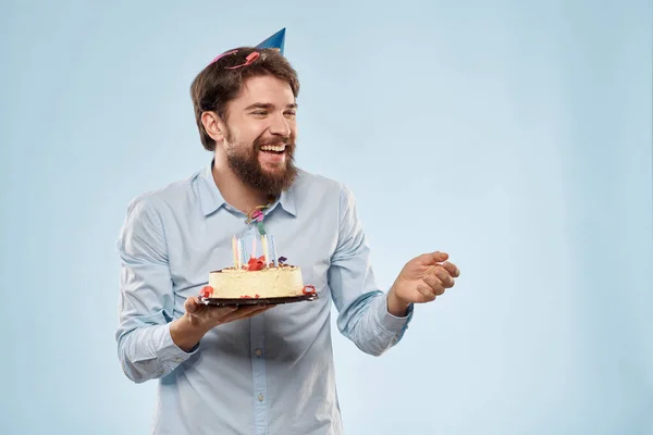 Skäggig man med en tallrik tårta på blå bakgrund och en födelsedagshatt på huvudet — Stockfoto