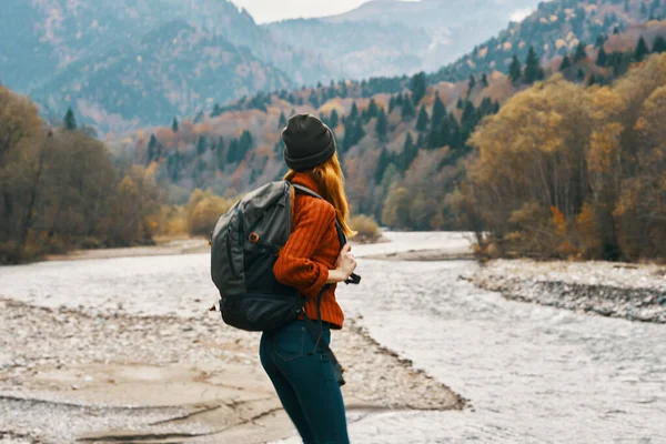 Donna in maglione con uno zaino riposo in montagna vicino al fiume in autunno nella natura e modello di alberi alti — Foto Stock