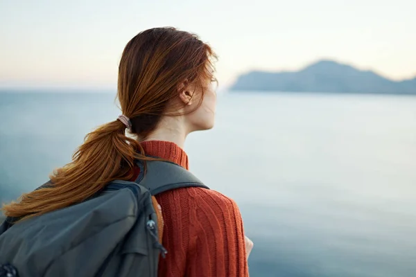 Donna turista in un maglione arancione e con uno zaino sulla schiena sta riposando vicino al mare e le montagne in lontananza — Foto Stock