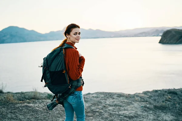 Щаслива жінка в червоному светрі з рюкзаком на спині відпочиває в природі біля моря і гір на відстані — стокове фото