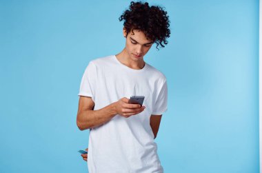Beyaz tişörtlü, kıvırcık saçlı, elinde telefon teknolojisi mavi arka plan.