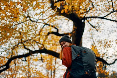 Kırmızı kazaklı bir sırt çantasıyla mutlu yolculuklar ve ağaçların yanındaki parkta kot pantolonlar.