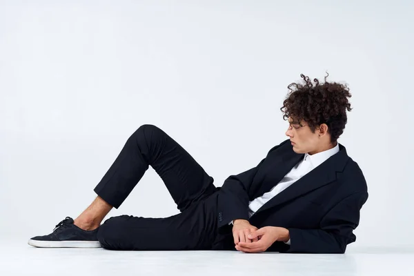 Ένας τύπος έφηβος με κοστούμι ξαπλώνει στο πάτωμα και αθλητικά σγουρά μαλλιά φως φόντο — Φωτογραφία Αρχείου