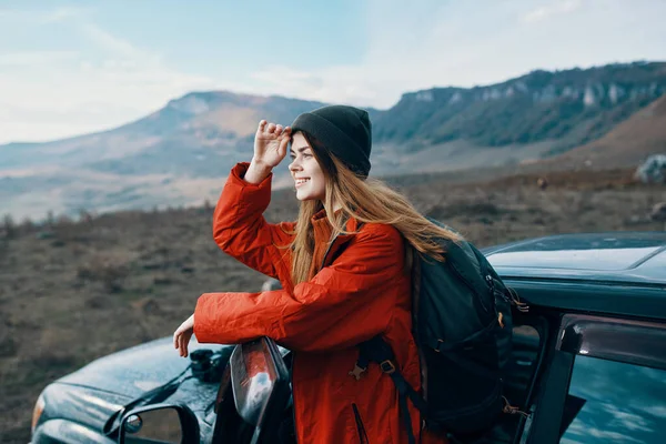 Kvinna i hatt med ryggsäck på ryggen lutad på dörren till en bil i bergen utomhus — Stockfoto